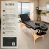 A black massage table is shown in a bright, modern room with a large window overlooking the ocean. Produktbild-Vorschau