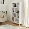 A white bookshelf with wooden legs is filled with books and decorative items, placed in a room with a sofa and a rug. Produktbild-Vorschau