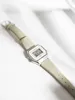 A digital watch with a beige strap is placed on a white surface with soft shadows. Produktbild-Vorschau