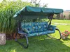 A garden swing with cushions is placed on a lawn in front of tall green plants. Podgląd zdjęcia produktu