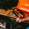 A person is placing an orange and black survival kit into a clear bag on a wooden shelf. Produktbild-Vorschau