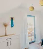 A bright, airy room with a slanted ceiling, featuring a white cabinet, a decorative angel figurine, dried grass in a vase, and sheer white curtains covering a glass door leading to an outdoor balcony. Produktbild-Vorschau