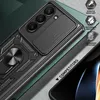 A close-up of a black phone case with a gold ring stand and a sliding camera cover, placed on a textured gray surface with a green background. Produktbild-Vorschau