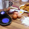 A digital kitchen scale with a stainless steel bowl, eggs, flour, and baked goods on a wooden table. Produktbild-Vorschau