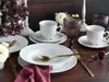 A beautifully set table with white porcelain dishes, gold cutlery, and purple glassware, adorned with flowers and a decorative candle holder. Produktbild-Vorschau