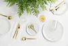A table setting with white plates, gold cutlery, a wine glass, and greenery. Voir les images du produit