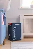 A navy laundry basket with the word "Laundry." printed on it in white is placed on a wooden floor next to a white radiator and a blue cabinet. Towels are visible inside the laundry basket and on a shelf of the cabinet. Náhled obrázku produktu