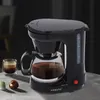 A black coffee maker with a glass carafe filled with coffee sits on a table, with a glass of coffee and two coffee pods next to it. Produktbild-Vorschau