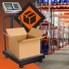 A digital scale with a cardboard box on it, set against a backdrop of a warehouse with shelves of products. Voir les images du produit