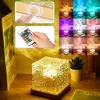 A collage of images showing a cube-shaped lamp with a textured surface, a remote control, and various color light projections. The main image shows the lamp on a wooden surface with a plant and a book. Produktbild-Vorschau