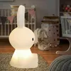 A white bunny-shaped LED night light is placed on a rug in a child's room, with a crib and storage baskets in the background. Produktbild-Vorschau