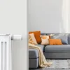 A smart radiator thermostat attached to a white radiator, with a grey sofa and decorative pillows in the background. Náhľad obrázku produktu