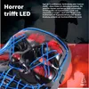 A close-up of a black LED mask with blue and red accents, with a dark, silhouetted background featuring figures and pumpkins. Produktbild-Vorschau