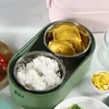 A close-up view of a green electric lunchbox with two stainless steel compartments, one filled with white rice and the other with fried food. Produktbild-Vorschau