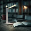 A white LED desk lamp illuminates an open book on a wooden desk, with bookshelves in the blurred background. Produktbild-Vorschau