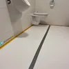 A clean, modern restroom with a urinal and toilet, featuring a white floor with a gray line and yellow border. Produktbild-Vorschau