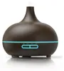 A dark brown aroma diffuser with a wood grain finish, emitting a visible mist from the top. It has a blue LED light strip around its base with illuminated buttons. Anteprima dell’immagine prodotto