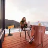 A woman sits on a deck in a snowy mountain setting, holding a mug and enjoying the view, with a patio heater nearby. Anteprima dell’immagine prodotto