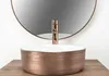 A rose gold basin sink with a copper faucet is placed on a wooden surface in front of a round mirror. Produktbild-Vorschau
