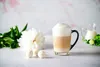 A glass mug of latte macchiato with foam, served with white candies and a white peony flower. Podgląd zdjęcia produktu