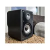 A black bookshelf speaker sits on a wooden table in a living room setting. Produktbild-Vorschau