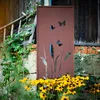 A decorative metal screen with butterfly and plant motifs stands in a garden, surrounded by flowers and greenery, next to a rustic wooden building. Náhled obrázku produktu