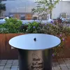 A round metal table with a black lid sits in an outdoor patio area with plants and seating in the background. Podgląd zdjęcia produktu