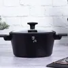 A black cooking pot with a glass lid sits on a wooden surface in front of a white brick wall. Podgląd zdjęcia produktu