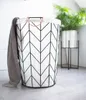 A laundry basket with a geometric pattern and a towel draped over it, placed on a tiled floor next to a potted plant. Produktbild-Vorschau