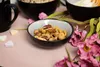 A close-up of a bowl of mixed nuts, with decorative flowers and heart-shaped candies on a pink surface. Náhled obrázku produktu