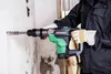 A person wearing gloves is using a Hikoki rotary hammer drill to drill into a wall. Produktbild-Vorschau