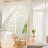 A bright, airy room with sheer white curtains blowing in the breeze, a wicker chair, and a small table with flowers. Produktbild-Vorschau