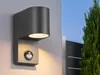 A modern outdoor wall light with a motion sensor is mounted on a gray wall next to a black gate and a green tree. Produktbild-Vorschau