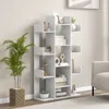 A white tree-shaped bookshelf filled with decorative items and books, placed in a room with a plant and a sofa visible. Podgląd zdjęcia produktu