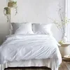 A neatly made bed with white bedding, two pillows, and a duvet, set against a textured white wall. Two hanging plants are visible in the upper left corner, and a vase with dried flowers and a teacup are on a side table to the right. Produktbild-Vorschau