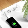 A white power strip with two AC outlets and two USB ports is shown on a white surface, with a smartphone plugged into one of the USB ports and charging. A plant is visible in the background. Podgląd zdjęcia produktu