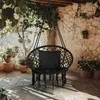 A black macrame hanging chair with a black cushion is set up on a stone patio under a wooden overhang with greenery. Voir les images du produit
