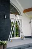 A multi-purpose ladder is set up in an A-frame configuration on a paved patio in front of a large window and a dark stone wall. Produktbild-Vorschau