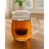 A glass tea mug with an infuser and lid, filled with tea, sits on a wooden table. Produktbild-Vorschau