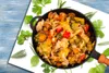 A close-up shot of a cooked vegetable dish in a black pan, surrounded by fresh herbs on a white surface and a blue wooden background. Náhľad obrázku produktu