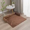 A brown quilted pet bed with raised edges is placed on a light wooden floor, with a vase of greenery and a small side table with books in the background. Náhled obrázku produktu