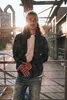 A man with long hair stands in an industrial setting, leaning on a railing. The sun is setting behind him, creating a warm glow and lens flare. Produktbild-Vorschau