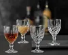 Four crystal liqueur glasses are arranged on a dark surface, with blurred bottles in the background. Podgląd zdjęcia produktu