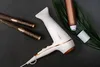 A white hairdryer with gold accents is laid out on a dark gray surface, alongside two hairbrushes and a bronze bottle with a white cap, partially covered by a large green leaf. Anteprima dell’immagine prodotto