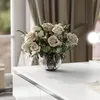 A bouquet of beige roses and greenery in a glass vase on a white surface, with a window and curtains in the background. Produktbild-Vorschau