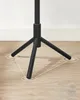The base of a black tripod stand is shown on a light brown wooden floor with white trim in the background. Náhled obrázku produktu