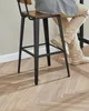 A person sitting on a black and wood bar stool with a herringbone wood floor. Produktbild-Vorschau