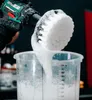 A person in black gloves is holding a drill with a brush attachment over a measuring cup filled with soapy water. Produktbild-Vorschau