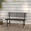 A dark metal garden bench sits on a wooden deck in front of a white paneled wall with some greenery hanging from above. Podgląd zdjęcia produktu