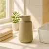 A beige ceramic soap dispenser sits on a white countertop next to a small potted plant and folded towels, with a window in the background. Podgląd zdjęcia produktu